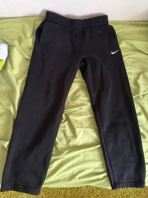 Svarta mjukisbyxor från Nike - Säljer ett par svarta mjukisbyxor från Nike med klassisk vit logga på vänster lår. Byxorna har resår i midjan och smal passform. Perfekta för chill eller träning. Mjukt material och fickor på sidorna.