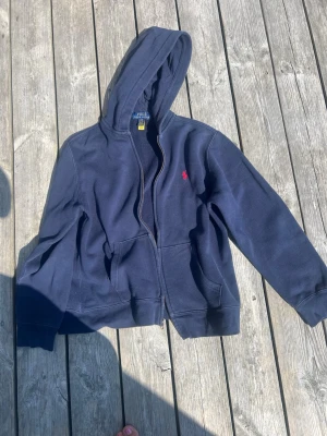 Mörkblå hoodie med dragkedja, stl 14/16y från Polo Ralph Lauren - Säljer en mörkblå hoodie, från Polo Ralph Lauren, med dragkedja och huva. Hoodien har två fickor framtill och ett litet rött broderat märke på bröstet. Perfekt för chill dagar och enkel att matcha med jeans eller mjukisbyxor.