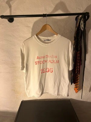 Vit Acne Studios t-shirt med tryck - Vit t-shirt från Acne Studios med rött tryck på bröstet där det står 'Acne Studios STOCKHOLM 1996'. Klassisk rund hals och korta ärmar. Perfekt för dig som gillar minimalistisk och stilren design. Dåligt croppad men ger den lite acne vibe ändå! 