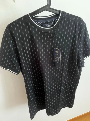 Svart mönstrad t-shirt Nickel & Dime - Svart t-shirt från Nickel & Dime med diskret grått mönster över hela tröjan. T-shirten har rund halsringning med vit kant och korta ärmar med vit detalj vid ärmslut. Tillverkad i mjuk bomull för en skön känsla.