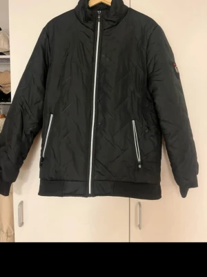 Svart pufferjacka med reflexdetaljer - Säljer en svart pufferjacka med coola reflexdetaljer längs dragkedjan och fickorna. Jackan har hög krage och ett märke på ena ärmen med texten 'Fashion G'. Perfekt för kalla vinterdagar och riktigt snygg streetstil.
