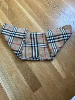 Beige rutig halsduk från Burberry - Klassisk beige halsduk från Burberry i 100% kashmir. Den har det ikoniska rutmönstret i svart, vitt och rött samt fransar i kanterna. Tillverkad i Skottland och supermjuk, perfekt för att lyfta vilken outfit som helst.