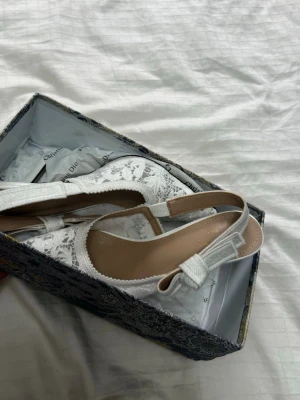 Vita spetsiga pumps med rosett - Säljer ett par eleganta vita pumps med spetsdetaljer och öppen häl. Skorna har en fin rosett på remmen och spetsig tå. Klacken är smal och ger en klassisk look. Perfekta för dig som vill ha något extra till outfiten.