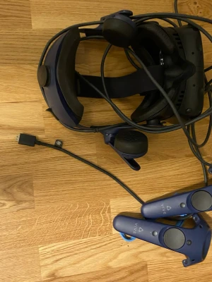 HTC Vive VR-headset med kontroller - Säljer ett HTC Vive VR-headset med två blå handkontroller och tillhörande kablar. Headsetet har inbyggda hörlurar och justerbart huvudband. Perfekt för att ta gamingupplevelsen till nästa nivå med VR. Allt är i snygg, modern design.