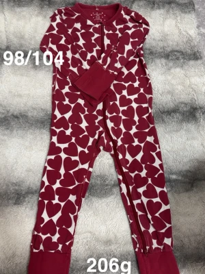 Rödvit pyjamas med hjärtmönster - Mysig pyjamas i röd och vit med stora hjärtan över hela plagget. Dragkedja framtill och muddar vid ärmar och benslut för extra komfort. Perfekt för sköna hemmakvällar eller sovstunder. 🌸 Små fläckar. Storlek 98/104 