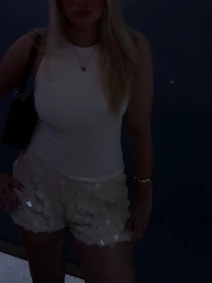 Vita paljettshorts med volang - Storlek L men passar M! Säljer ett par vita shorts med glittriga paljetter och volangdetaljer. Shortsen har en hög midja och är perfekta för dig som vill sticka ut lite extra. De är tillverkade i ett syntetmaterial som ger en skön känsla och snygg passform.
