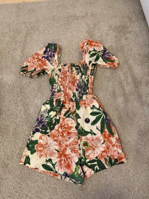 Blommig jumpsuit med puffärm - Byxdress från H&M med stora blommönster i rosa, lila och grönt. Jumpsuiten har puffärmar och smockad överdel som ger en snygg passform. Bomullsmaterialet är lätt och bekvämt. Kort modell med breda ben.