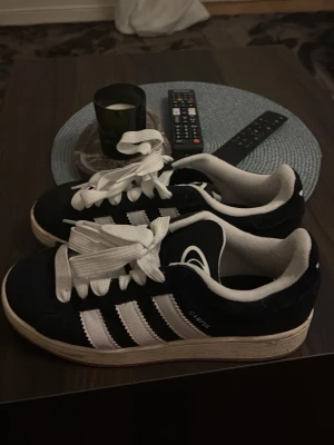 Adidas Campus svartvita sneakers - Säljer ett par klassiska Adidas Campus sneakers i svart med vita detaljer och breda vita snören. Skorna har tre vita ränder på sidan och Adidas-logga på tungan. Ovandelen är i mocka och sulan är vit. Perfekt för dig som gillar retrostil. Använd max 2 gånger. Sänka pris vid snabb affär