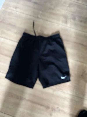 Svarta träningsshorts från Nike - Svarta träningsshorts från Nike med klassisk logga på ena benet. Shortsen har elastisk midja med snörning för bästa passform och är tillverkade i ett lätt och svalt material som passar perfekt för sport och träning.