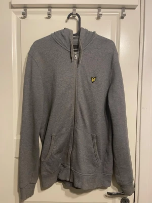 Grå hoodie med dragkedja från Lyle & Scott - Grå hoodie med dragkedja från Lyle & Scott. Klassisk modell med huva, två fickor framtill och gul broderad logga på bröstet. Mjuk bomullsblandning som är skön att ha på sig. Perfekt för en avslappnad stil.