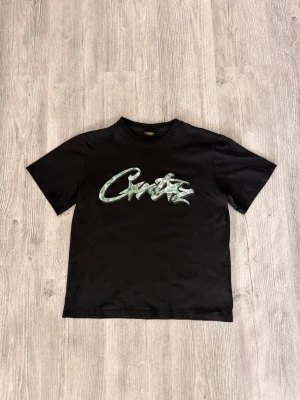 Svart t-shirt med grön graffiti-text - Snygg svart t-shirt från Crate med stor grön graffiti-inspirerad text på bröstet. Klassisk passform med rund hals och korta ärmar. Tillverkad i mjuk bomull som känns skön mot huden. Perfekt för dig som gillar streetwear och vill sticka ut.