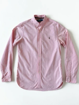 Rosa skjorta från Polo Ralph Lauren - Klassisk rosa skjorta från Polo Ralph Lauren med button-down krage och broderad logga på bröstet. Skjortan har långa ärmar, knappar framtill och är tillverkad i mjuk bomull. Perfekt för en stilren och avslappnad look.