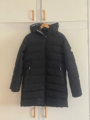 Svart lång dunjacka från Moncler - Säljer en svart lång dunjacka från Moncler med huva och dragkedja framtill. Jackan har två fickor med dragkedja och diskret Moncler-logga på ärmen. Klassisk quiltad design och varm vaddering, perfekt för kalla vinterdagar.