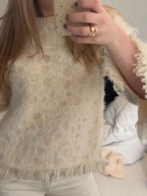 Beige blus med fransar och spets - Säljer en beige blus med unikt spetsmönster och fransar längs nederkant och ärmar. Blusen har korta ärmar och en rund halsringning, perfekt för dig som gillar detaljer och en lite bohemisk stil.