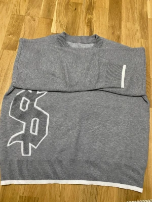 Grå tröja med vit dollartryck - Snygg grå tröja med stort vitt dollar-tecken på framsidan. Tröjan har ribbstickade muddar och nederkant med vit detalj. Perfekt för dig som gillar streetstyle och vill sticka ut lite extra.