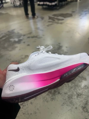 Nike vita sneakers med rosa sula - Snygga vita Nike sneakers med stickad ovandel och en fet rosa gradient på mellansulan. Skon har en mörkbrun yttersula med coolt mönster och klassisk Nike-logga på sidan. Perfekt för dig som vill sticka ut med stilrena men färgstarka skor.