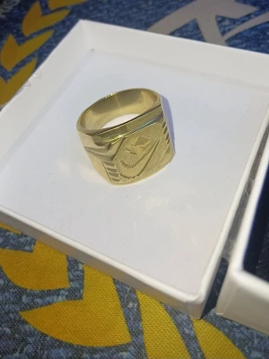 Nike klackring stämplad med "18k" & "750" från dödsbo  - Stilren guldfärgad ring med ingraverad Nike-logga och geometriska detaljer på sidorna. Ringen har en bred och platt design som ger ett coolt och sportigt intryck. Perfekt accessoar för dig som gillar streetwear och vill sticka ut. Obs. Den är stämplad som 18karats guld men det kan lika gärna vara av något annat material därav det billiga priset. Köp ej om du inte är införstådd med att det kanske inte är guld.