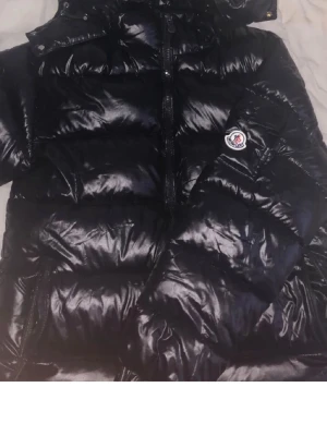 Svart dunjacka från Moncler - Snygg svart dunjacka från Moncler med glansig finish och klassisk logga på ärmen. Jackan har huva, dragkedja framtill och breda kanaler fyllda med dun som håller dig varm under kalla dagar. Perfekt för vintern och riktigt trendig stil.