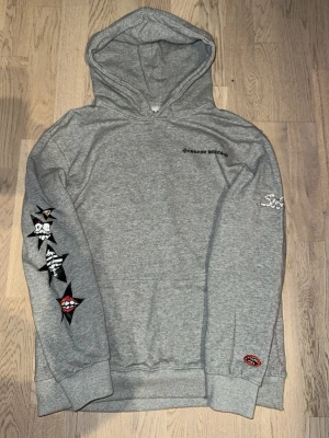Grå hoodie från Chrome Hearts - Grå hoodie från Chrome Hearts med coola tryck på ärmen och ryggen. På ryggen står det 'SUGGEST' och det finns stjärnor med olika motiv. Liten Chrome Hearts-logga på bröstet och ärmen. Perfekt för dig som gillar streetwear och unika detaljer. STORLEK S FAST PASSAR XS!!!