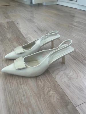 Beige slingback pumps med rosett - Aldrig använda stilrena vita/ beiga slingback-klackar med spetsig tå. Säljs endast pga fel storlek och har orginalförpavknibgen kvar. Skorna har öppen häl med justerbar rem och en smal klack. Tillverkade i ett glansigt material. 