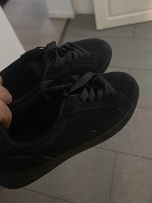 Helvita svarta sneakers med mockakänsla - Snygga helsvarta sneakers med låg profil och klassisk design. Skorna har ovandel i mockaliknande material och matchande svarta skosnören. Yttersulan är också svart och har ett diskret mönster för bra grepp. Perfekta för en clean och stilren look.