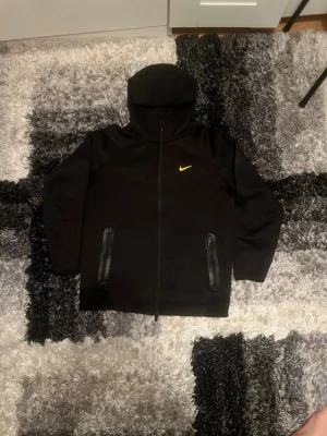 Svart Nike hoodie med gul logga - Säljer en svart hoodie från Nike med gul swoosh på bröstet och gul text på ryggen. Hoodien har dragkedja framtill, två fickor med dragkedja och en stor huva. Materialet är mjukt och bekvämt, perfekt för en avslappnad look. Obs de litet hål 