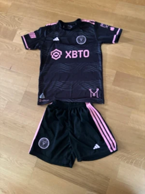 Inter Miami Messi matchställ svart/rosa - Snyggt Inter Miami matchställ med Messi 10-tryck. Svart tröja och shorts med rosa detaljer och loggor. Tröjan har klubbmärke, sponsortryck och Adidas-logga. Tillverkat i lätt och ventilerande material, perfekt för fotbollsträning eller match.