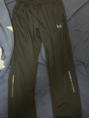 Svarta träningsbyxor Under Armour Reflex - Svarta träningsbyxor från Under Armour med färgglad logga på höften och reflexdetaljer med texten 'STORM' på benen. Byxorna har dragsko i midjan och är gjorda i ett lätt, sportigt material som passar perfekt för träning eller löpning.
