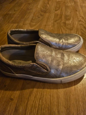 Guldiga slip-on sneakers - Snygga slip-on sneakers i metallic guld med reptilmönstrad yta och vit sula. Skorna har en låg profil och elastiska paneler på sidorna för enkel på- och avtagning. Perfekta för dig som vill sticka ut med en glansig touch till din outfit. Lite smuts på sulor men går säkert att få bort. Se bilder.