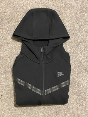 Svart NikeTech hoodie med dragkedja - Svart hoodie från Nike med hel dragkedja framtill och huva. Snygga grå reflexdetaljer i V-form över bröstet och klassisk Nike-logga på vänster sida. Tillverkad i mjukt material som passar perfekt för en sportig och avslappnad stil.