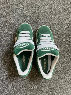 Gröna Adidas campus i mocka - Snygga gröna Adidas sneakers i mocka med vita detaljer och klassiska tre ränder på sidorna. Skorna har vit sula, vita skosnören och Adidas logga på plösen. Perfekta för dig som gillar retrostil och streetwear.