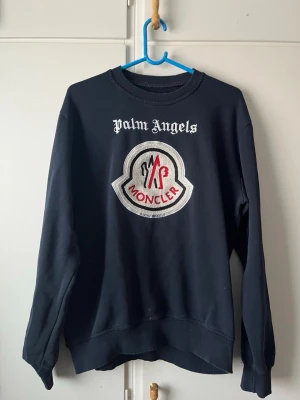 Palm Angels x Moncler svart sweatshirt - Svart sweatshirt från Palm Angels i samarbete med Moncler. Stor broderad Moncler-logga i vitt, rött och svart på bröstet samt Palm Angels-tryck ovanför. Tillverkad i 100% bomull och har ribbade muddar vid ärmslut och nederkant. Perfekt för en streetwear-look.