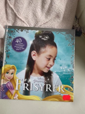 Disney Prinsessor Frisyrer bok - En färgglad bok med titeln 'Disney Prinsessor Frisyrer' som visar 40 olika prinsessfrisyrer med steg-för-steg-bilder. Omslaget har en turkos bakgrund, silverdetaljer och illustrationer av prinsessor. Perfekt för dig som gillar hårstyling och Disney.