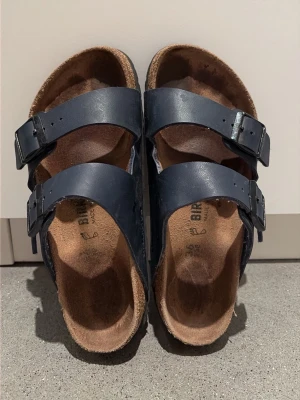Mörkblå Birkenstock sandaler - Säljer ett par klassiska mörkblå Birkenstock sandaler med två justerbara remmar i läder och metallspännen. Fotbädden är i kork och mocka för extra komfort. Perfekta för en avslappnad och trendig stil. I storlek 36, men jag som vanligtvis har 37 i skor passar perfekt i dessa. Var uppmärksam att storleken kan skiftas. 