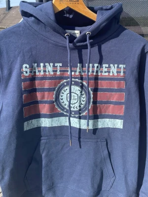 Blå hoodie från Saint Laurent - Snygg marinblå hoodie från Saint Laurent med tryck framtill i vitt och rött samt logga. Klassisk känguruficka och justerbar huva med snören. Perfekt för en avslappnad och trendig look. Mjuk bomullskänsla och streetwear-vibe.