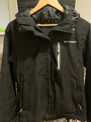 Svart vindjacka från Everest - Svart vindjacka från Everest med dragkedja med vita detaljer på. Jackan har minimalistisk design och är tillverkad i ett slitstarkt material som står emot vind. Perfekt för utomhusaktiviteter när du vill hålla stilen. Byten funkar också!