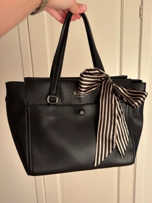 Söt väska från kate of london - Jättesöt väska från kate of london. Knappt använd och inga defekter. Det ingår även ett längre band så att man kan ha den som en crossbody. Mycket rymlig och har en söt liten rosett som man kan ta av💞 Skriv vid eventuella frågor!