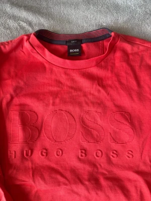 Röd sweatshirt från Hugo Boss - Snygg röd sweatshirt från Hugo Boss med stort broderat BOSS-logo på bröstet. Tröjan har rund halsringning, ribbade muddar och är tillverkad i mjuk bomull. Perfekt för en avslappnad och trendig stil.