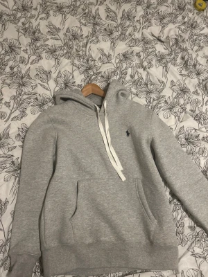 Grå hoodie från Polo Ralph Lauren - Säljer en stilren grå hoodie från Polo Ralph Lauren med klassisk huva och vita dragsnören. Tröjan har en broderad logga på bröstet och en stor magficka. Tillverkad i mjukt sweatshirtmaterial som är perfekt för chill dagar.
