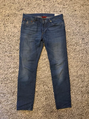  Brunello Cucinelli Jeans - Säljer ett par blå slim fit jeans från Brunello Cucinelli med klassisk femficksmodell och snygga slitningar. Jeansen har en diskret logga på myntfickan och är tillverkade i mjuk bomull. Perfekta till vardagslooken!