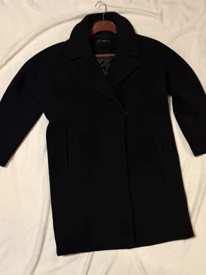 Svart ullkappa från Hallhuber - Stilren svart kappa från Hallhuber med klassisk krage och diskreta sidofickor. Kappan har rak passform och är fodrad med svart tyg på insidan. Perfekt för kyliga dagar och ger en snygg siluett till din outfit.