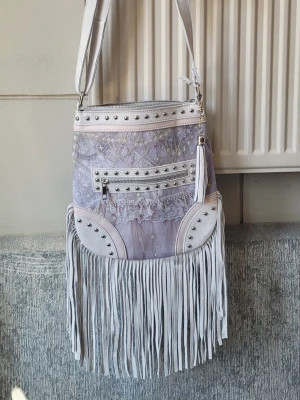 Ljuslila axelväska med fransar van asch boho  - Underbar väska från Van Asch i fint "spetstryck", med mockadetaljer Reglerbar axelrem, stängning med dragkedja, flera fickor, varav en utanpå med dragkedja. Ca mått 28 x 29 x 2 utan fransarna