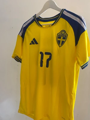 Sverige fotbollströja Adidas #17 Gyökeres - Gul och blå fotbollströja från Adidas med Sveriges landslagsmärke på bröstet och nummer 17 samt namnet Gyökeres på ryggen. Tröjan har korta ärmar, blå detaljer på axlarna och är tillverkad i ett lätt, ventilerande material perfekt för match och en stundande vm💛💙