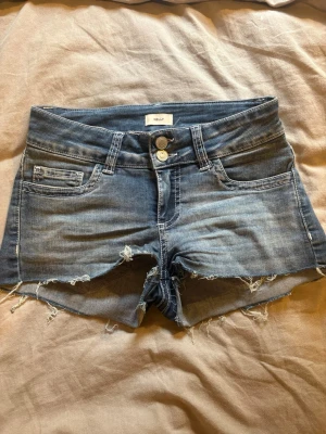 Blå jeansshorts från Nelly - Snygga blå jeansshorts från Nelly med råa kanter och slitna detaljer. Klassisk femficksmodell med dubbla knappar framtill och broderade bakfickor. Perfekta för varma dagar och en avslappnad stil.