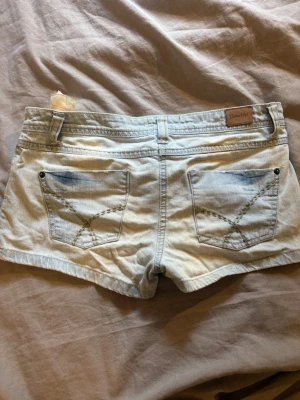 Ljusblå denimshorts från Denim Life - Snygga ljusblå denimshorts från Denim Life med klassisk femficksdesign och coola detaljer på bakfickorna. Perfekta för varma dagar och enkla att matcha med allt. Shortsen har bälteshällor och knappstängning framtill.