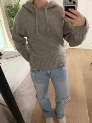 Grå stickad hoodie med huva - Mysig grå stickad hoodie med huva och dragsnören. Tröjan har ribbade muddar vid ärmslut och nederkant, vilket ger en snygg och avslappnad look. Perfekt för dig som gillar en enkel och stilren stil.