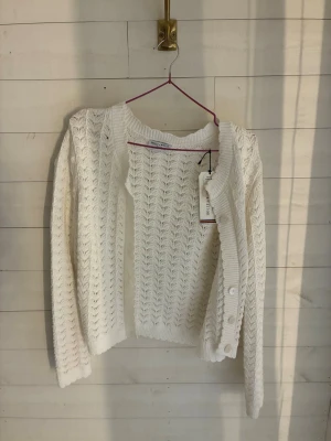 Vit stickad kofta med mönster - Säljer en vit stickad kofta med snyggt hålmönster och ribbade kanter. Koftan har långa ärmar och knappar framtill. Perfekt att slänga över en topp för en chill look. Materialet känns mjukt och luftigt. Knappen längst upp saknas men den följer med på sidan💗
