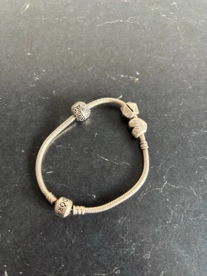 Pandora armband i silver med berlocker - Snyggt armband från Pandora i äkta silver med flera unika berlocker, bland annat hjärta och mönstrad kula. Armbandet har klassisk ormkedja och detaljerade pärlor som ger en personlig touch. Perfekt accessoar för att uttrycka din stil.