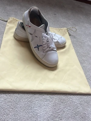 Vita Axel Arigato sneakers med fågelmotiv - Snygga vita sneakers från Axel Arigato med broderade fåglar i olika färger på sidorna. Skorna har klassisk låg modell, vita skosnören och är tillverkade i läder. Perfekta för dig som vill ha en stilren men unik look.