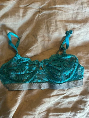 Turkos spets-bh med bred resår - Snygg turkos bh i spets med bred grå resår under bysten. Justerbara axelband och hakknäppning bak. Bh:n har ett feminint och trendigt utseende med transparent spetsmaterial och dekorativa detaljer.